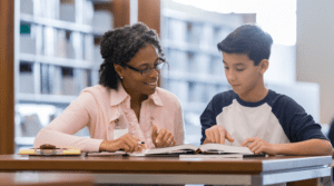 A student and homeschool tutor sit in a library working on assignments.